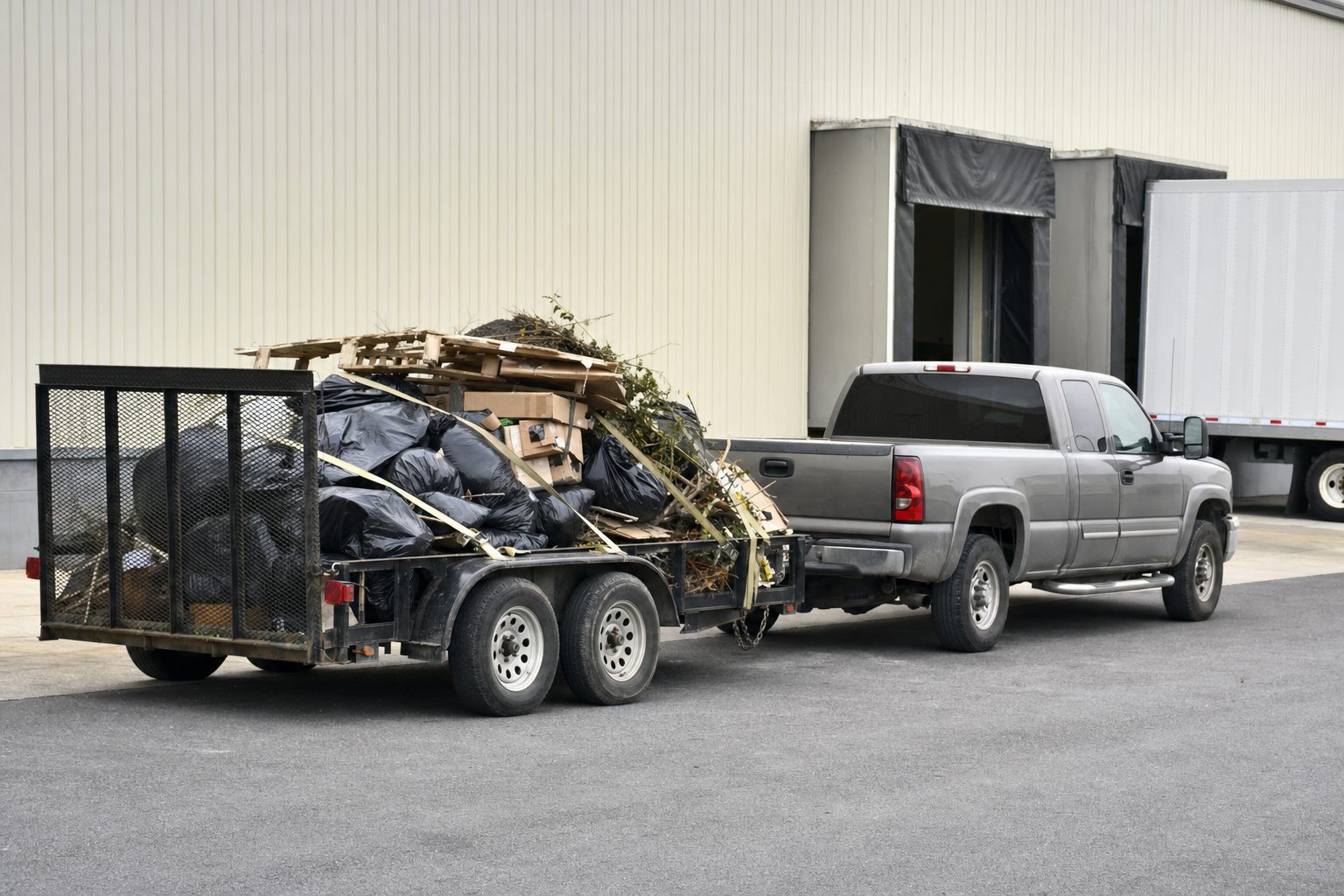 A pickup truck and trailer loaded with bagged trash, pallets, and debris at an industrial loading dock